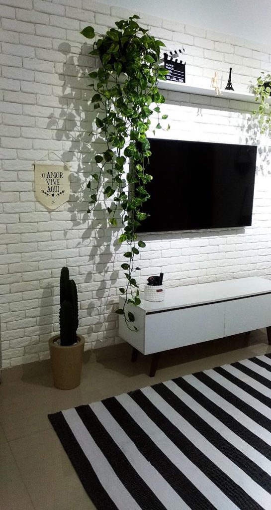 Decoração com plantas da sala de estar de cliente Madesa. O ambiente conta com um cacto e uma planta pendente, além do rack Reims, tapete listrado e parede em tijolinhos brancos.
