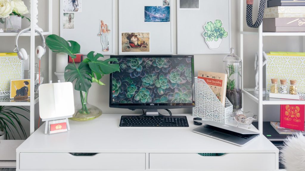 Escritório em casa equipado com escrivaninha branca e um computador preto. Na tela do monitor há imagens de plantas.