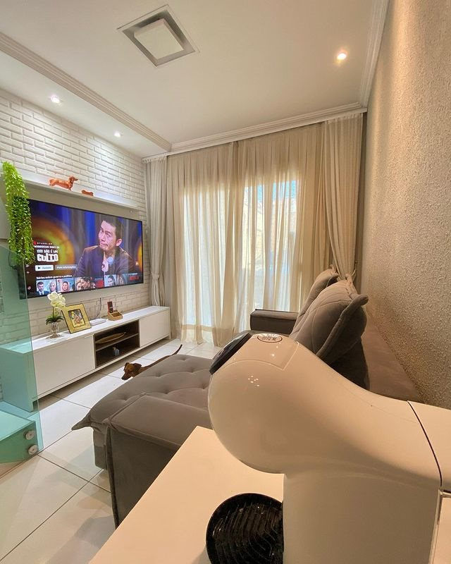 A imagem ilustra uma sala de estar com uma tv instalada em cima de um hack branco, representando um presente para casa nova.