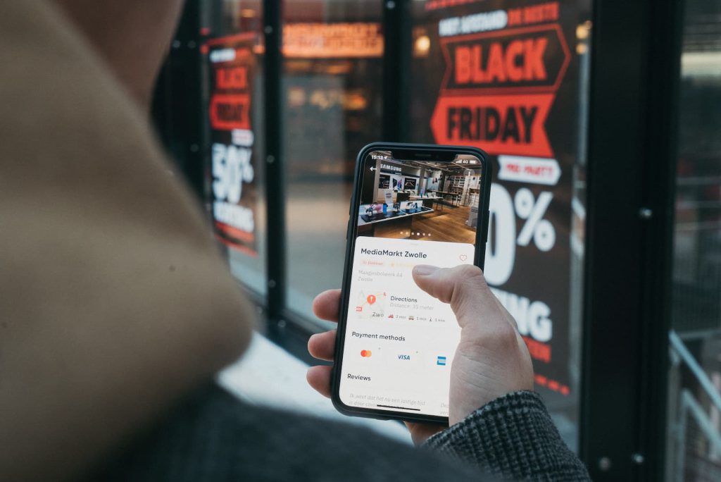 Pessoa conferindo promoções de Black Friday no celular em frente a uma vitrine com descontos anunciados em adesivos vermelhos e brancos.