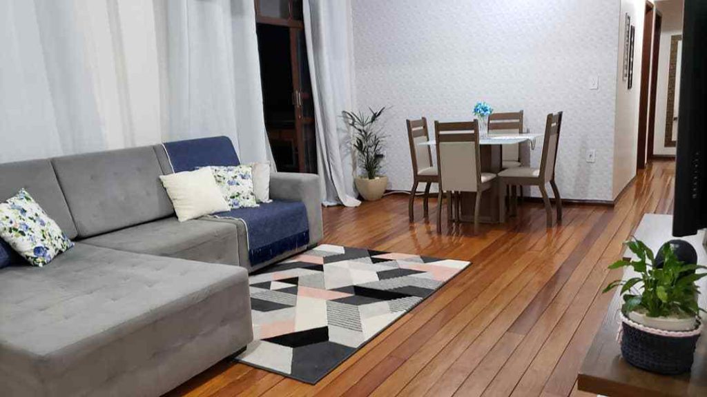 Mesa de jantar quadrada ao canto direito, um sofá com almofadas e tapetes compondo a imagem de uma sala de estar com uma sala de jantar no mesmo ambiente. 