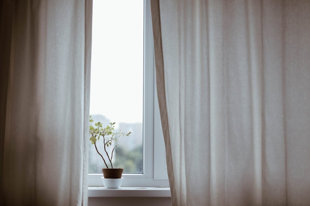 Janela com cortina em cor clara e um vaso de plantas como exemplo dos tipos de cortina