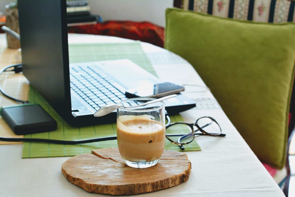 Mesa com um notebook, um par de óculos e um copo de café em cima de um suporte de madeira como exemplo de decoração de home office.