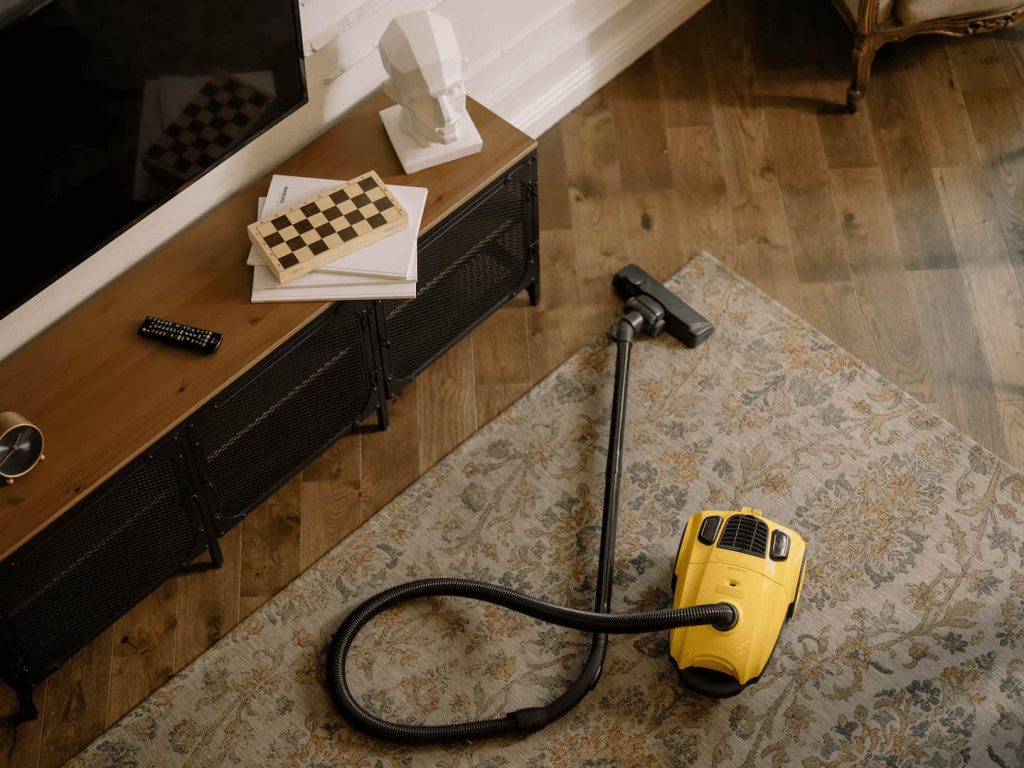 Aspirador de pó amarelo em cima do tapete da sala, que também contem um rack preto, uma TV e objetos decorativos.