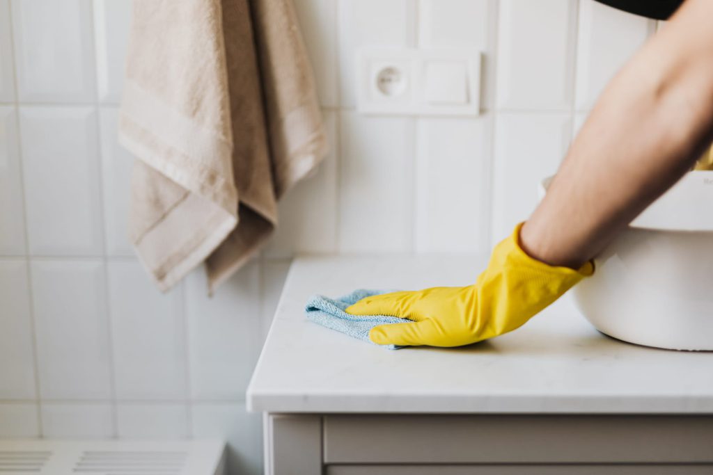 Imagem de uma pessoa limpando a pia do banheiro, usando luvas amarelas e um pano azul, como exemplo da limpeza de casa.