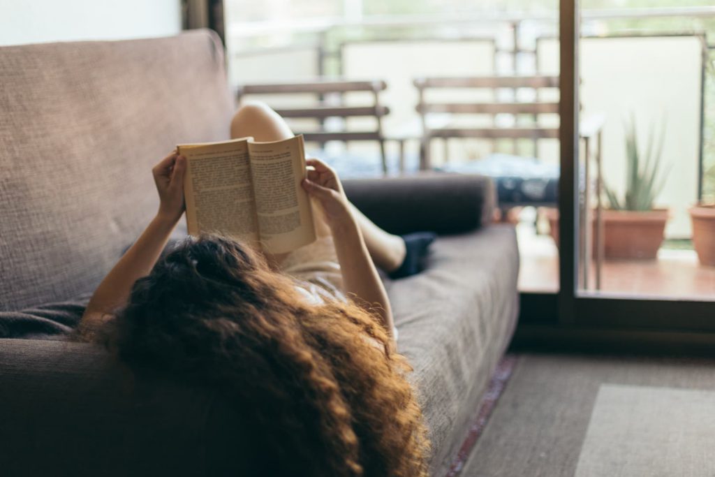 Mulher lendo um livro deitada em um sofá como exemplo de como é morar sozinho