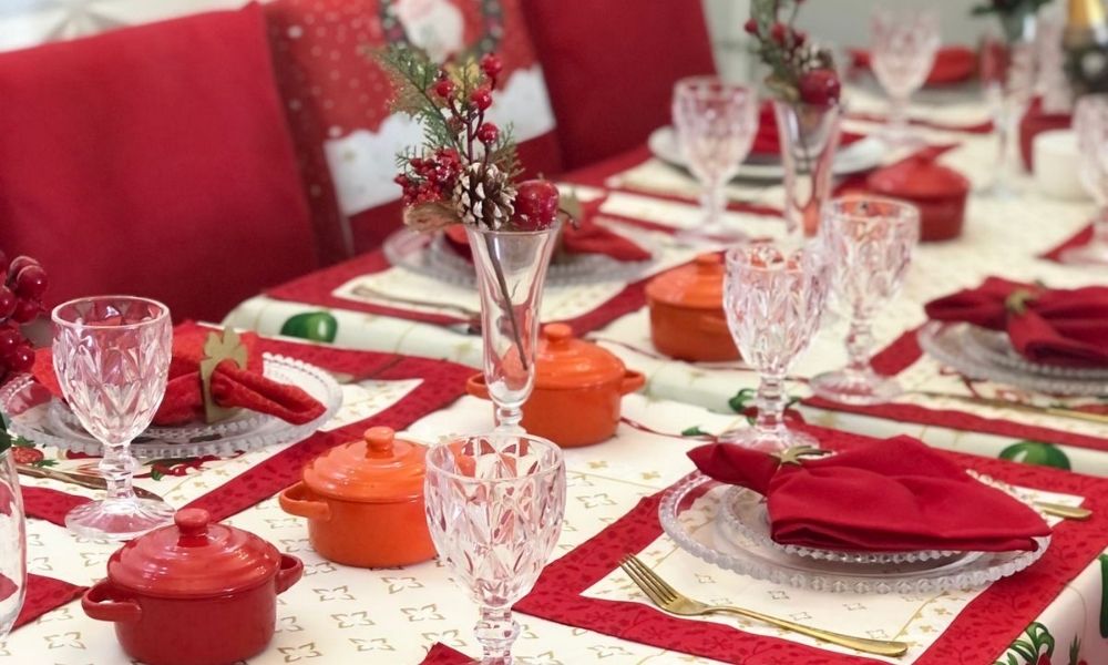 Mesa posta de natal com guardanapos vermelhos, taças de vidro e enfeites natalinos.