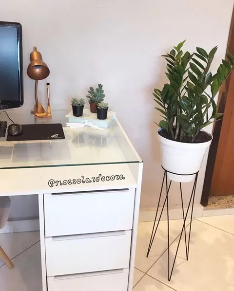 A imagem ilustra uma escrivaninha branca com um computador e um vaso de plantas ao lado, demonstrando ideias para trabalhar em casa.