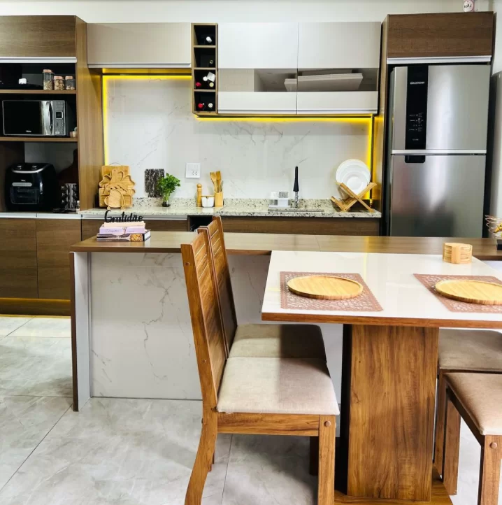 A imagem ilustra uma cozinha com armários de madeira, utensílios domésticos e uma mesa com cadeiras e pratos sobre ela, na tentativa de mostrar como organizar armário de cozinha.