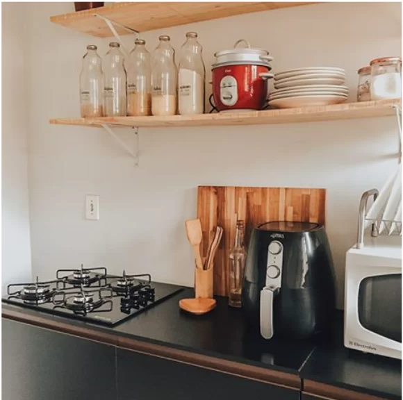 A imagem ilustra como organizar armário de cozinha mostrando um cooktop preto em uma bancada preta composta por itens de madeira, uma airfryer e uma prateleira com temperos.