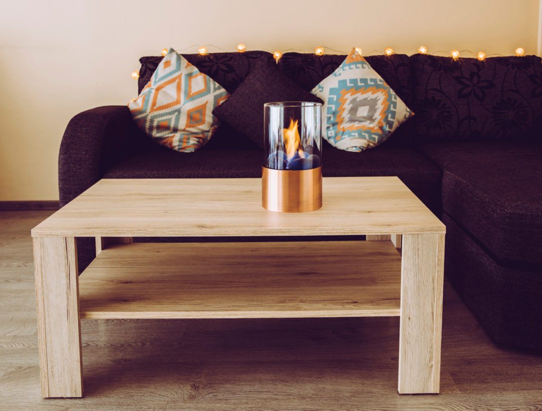A imagem ilustra uma sala com lareira ecológica, composta por um sofá com almofadas coloridas e uma mesa de madeira de cor clara.
