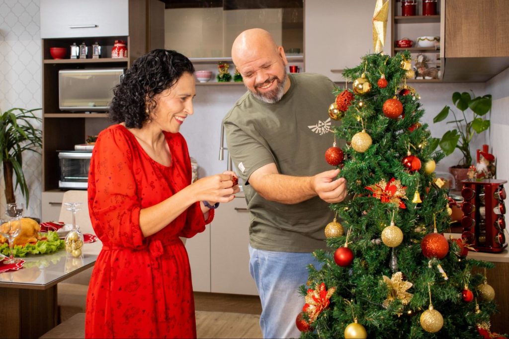 Casal de adultos, decorando juntos uma árvore de Natal, sorrindo. Ao fundo, uma cozinha Madesa e uma mesa de jantar, ambos decorados.