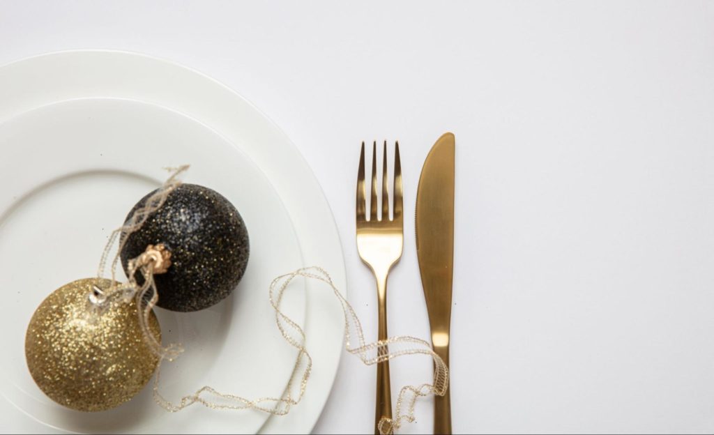A imagem ilustra um prato branco com talheres dourados ao lado e uma bola de decoração de fim de ano simbolizando o que pode ser uma decoração para ceia de ano novo simples e barata.