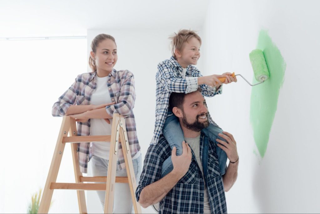 Uma família composta por um homem, uma mulher e uma criança, pintando de verde uma parede de casa