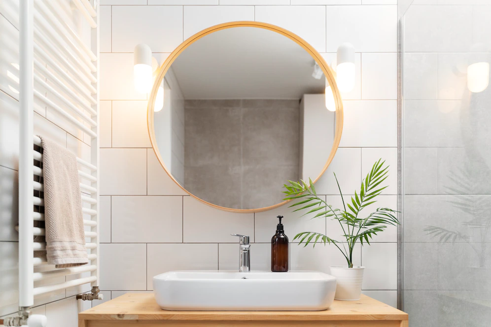 A imagem mostra um espelho redondo com detalhes em madeira e uma pia branca em um banheiro clean.