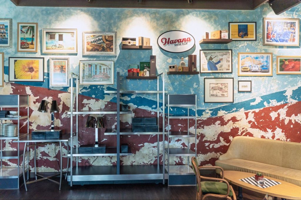 A imagem ilustra uma parede azul com vários quadros e posters, além de estantes em metal, decorando uma garagem simples.