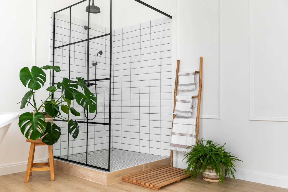 A imagem mostra um banheiro de paredes brancas e box de vidro em detalhes pretos com plantas, representando a estética de um banheiro clean.