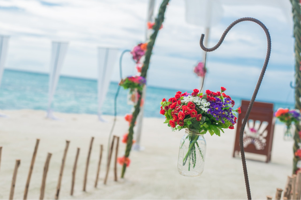 A imagem ilustra um vaso de flores coloridas pendurado a um suporte de ferro, colocando dentro de um vidro transparente, representando a decoração de casamento na praia.