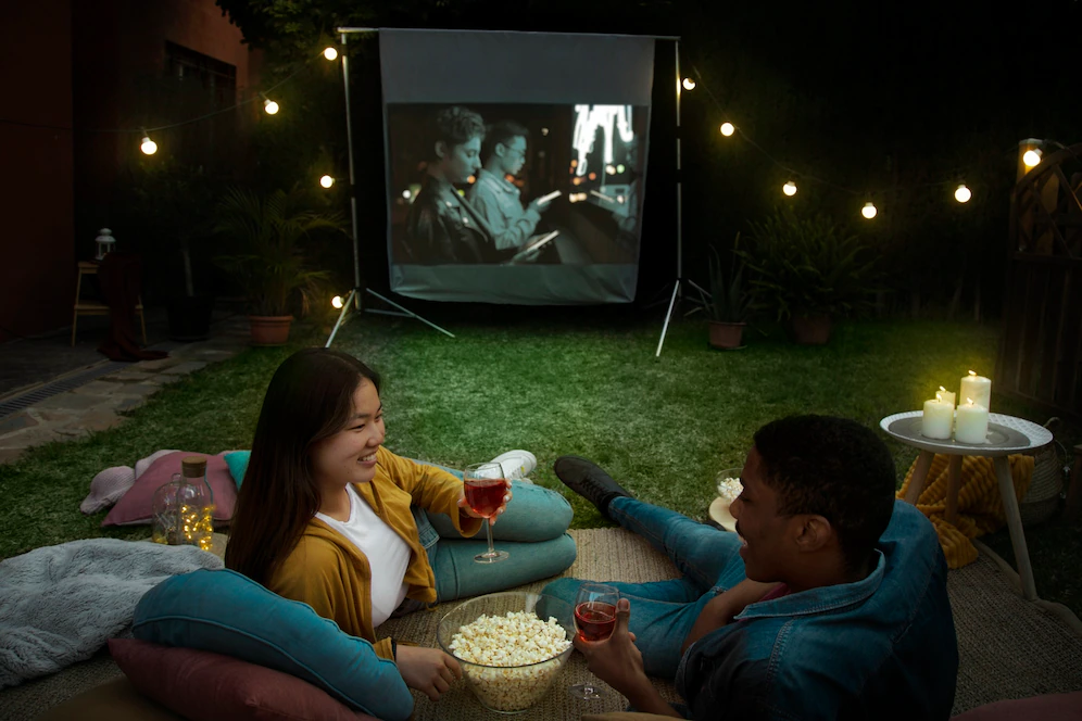 A imagem ilustra um casal deitado em um quintal com grama e luzes a noite, olhando um para o outro enquanto um projetor completa a sala de cinema residencial no quintal.
