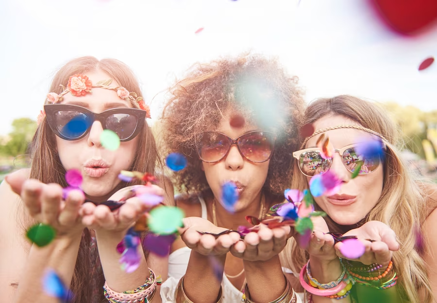 A imagem ilustra três mulheres jovens soprando confetes fantasiadas com acessórios na sua festa tema carnaval.