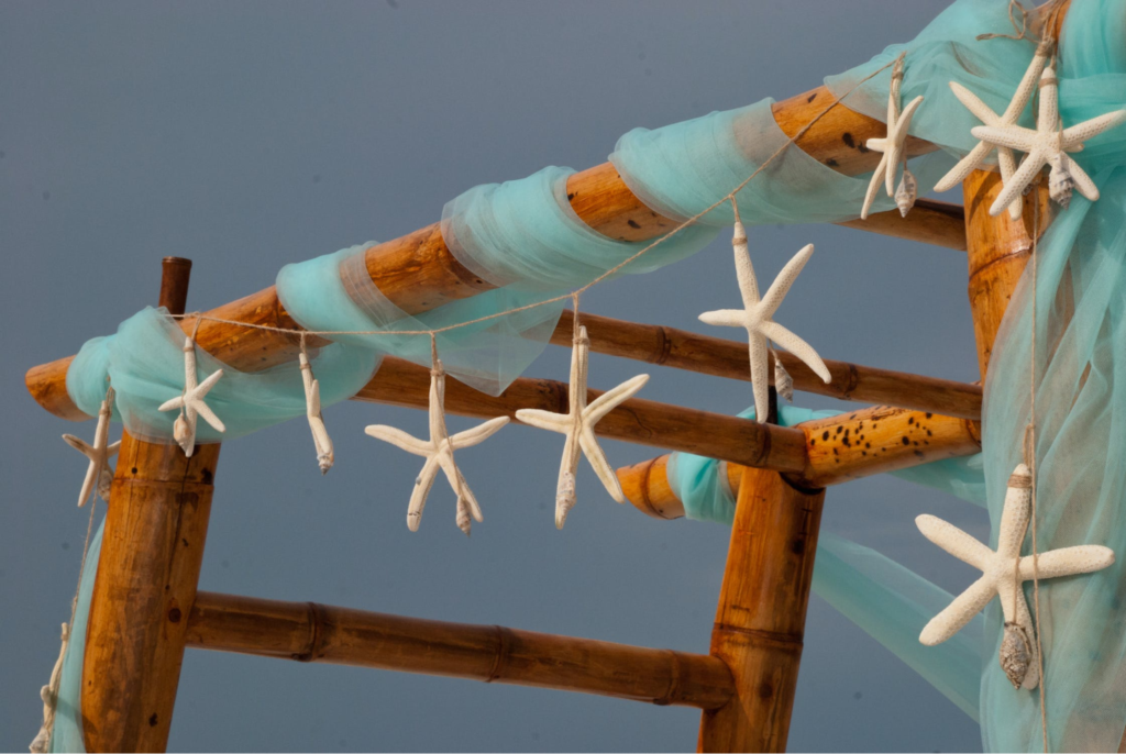 A imagem ilustra tecidos azul tifanny enrolados em um gazebo de madeira compondo uma decoração de casamento na praia.