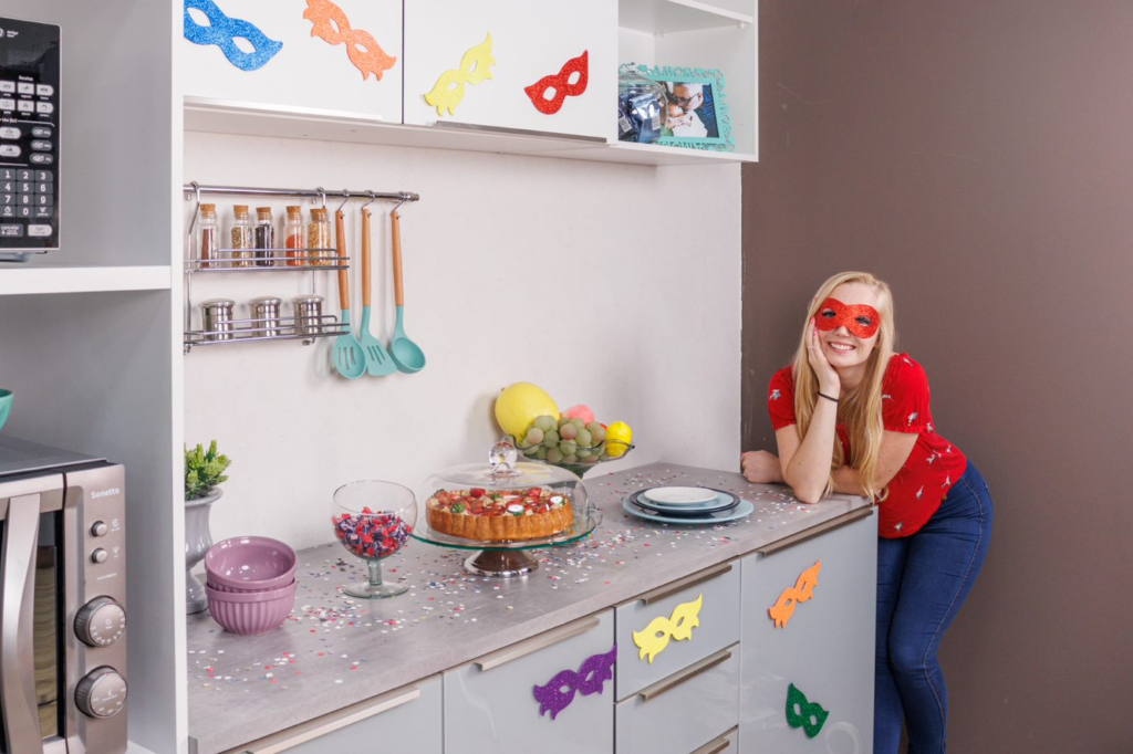 A imagem ilustra uma mulher jovem e loira de cabelos longos em sua cozinha decorada com tema festa carnaval.