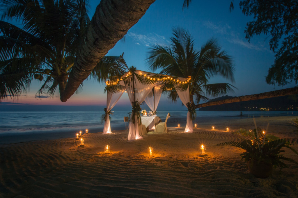 A imagem ilustra uma decoração de casamento na praia à noite, à luz de velas.