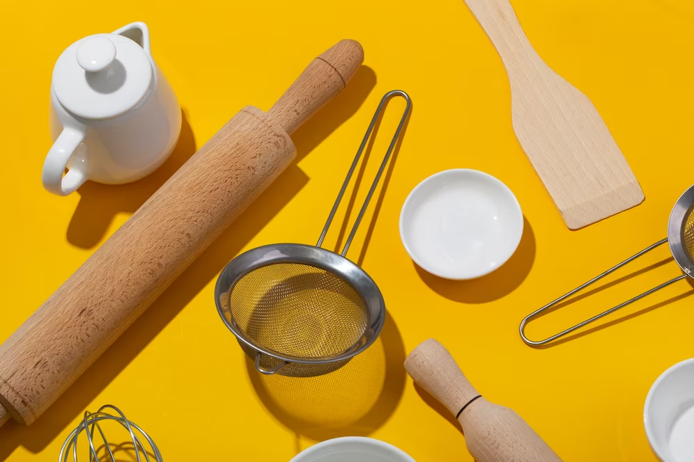 A imagem ilustra uma lista de utensílios de cozinha num fundo amarelo, como por exemplo, um amassador de pão, uma peneira e um bule.