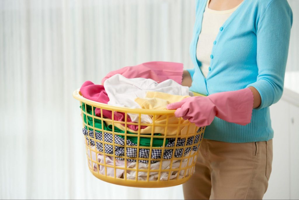 A imagem ilustra uma mulher segurando um cesto de roupa suja, simbolizando os afazeres domésticos.