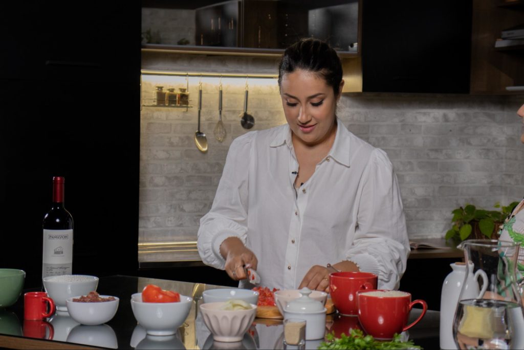 A imagem ilustra uma mulher branca de cabelos curtos pretos e blusa branca preparando refeições, simbolizando a rotina de afazeres domésticos.