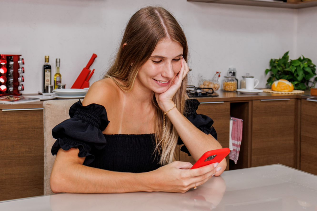 A imagem ilustra uma mulher loira de blusa preta segurando um celular vermelho na frente de uma cozinha planejada pequena, simples e barata.