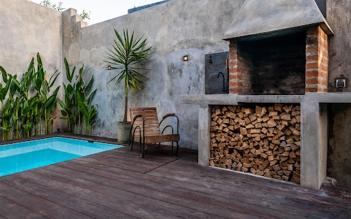 na imagem, vemos a área externa de uma residência, que conta com uma piscina, uma churrasqueira que leva lenha e um piso de madeira