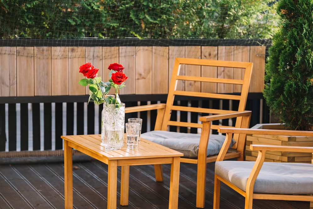 na imagem, vemos duas cadeiras de madeira que estão próximas de uma mesinha de centro, levando um vaso de flores vermelhas. Todas estão sobre um deck de madeira na área externa de uma casa.