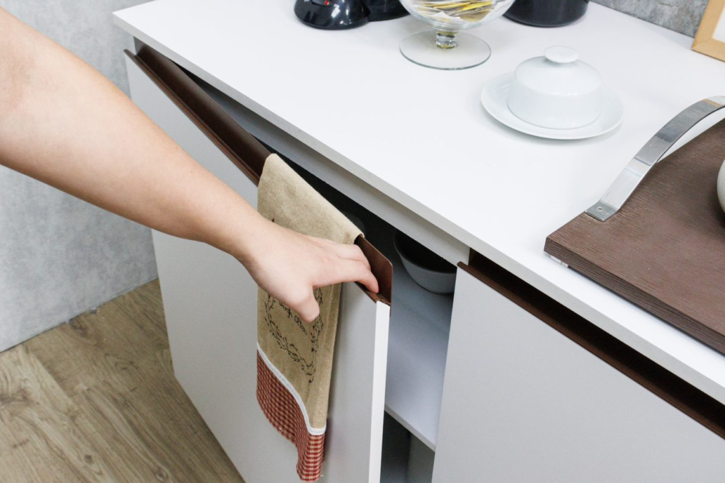 na imagem, uma mão feminina está abrindo um armário de cozinha Madesa na cor branca, com uma bancada e alguns objetos de cozinha em cima dela