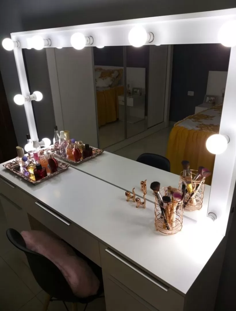 Penteadeira Madesa, na cor branca, com espelho e luzes de LED. Em cima, pincéis de maquiagem e uma bandeija com perfumes e outros produtos de beleza.