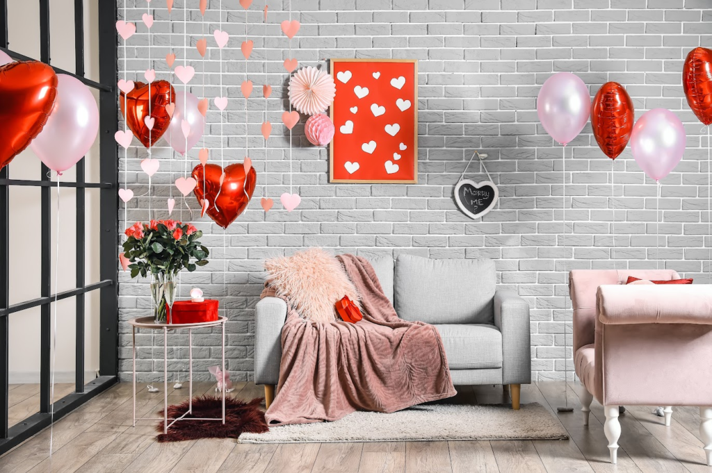 A imagem ilustra uma sala decorada com balões vermelhos em forma de coração, um sofá cinza e flores, simbolizando ideias para o dia dos namorados.
