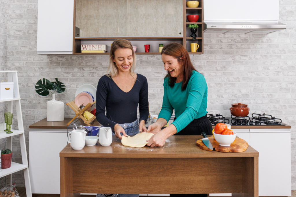 A imagem ilustra mãe e filha cozinhando uma massa em uma cozinha branca, representando o presente para o Dia das Mães.