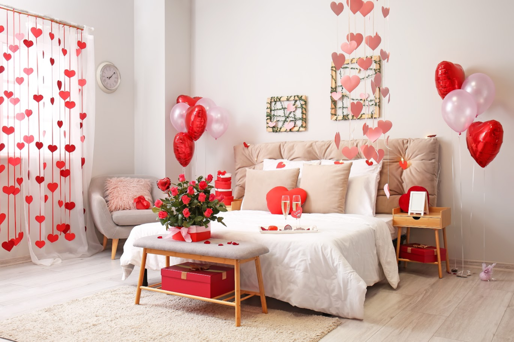 A imagem ilustra um quarto decorado com ideias para o dia dos namorados. Tem uma cama ao centro e muitos detalhes em vermelho, como rosas, balões, etc.