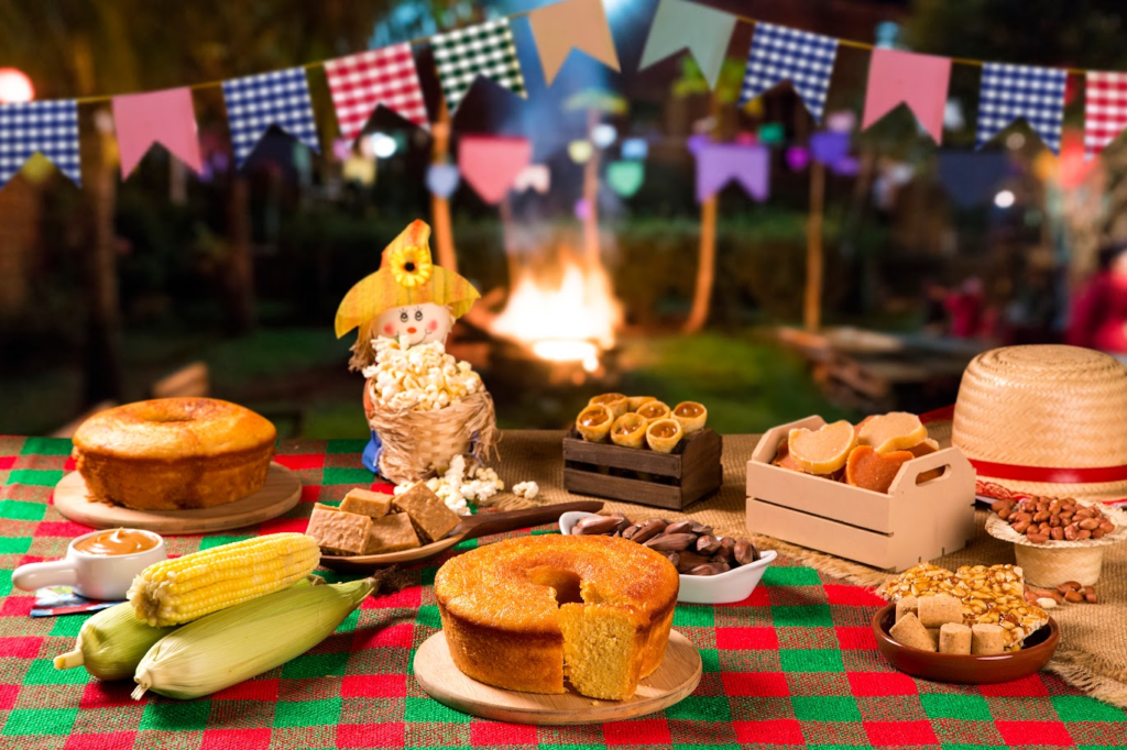 A imagem ilustra uma mesa decorada com o que tem na festa junina de mais tradicional em comidas. Ao fundo há uma fogueira e bandeirinhas.