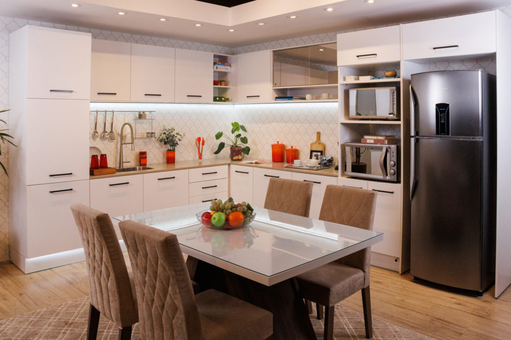 A imagem ilustra uma cozinha planejada em L Madesa branca, cm eletrodomésticos e uma mesa de jantar ao centro. Essa mesa tem uma fruteira colorida.