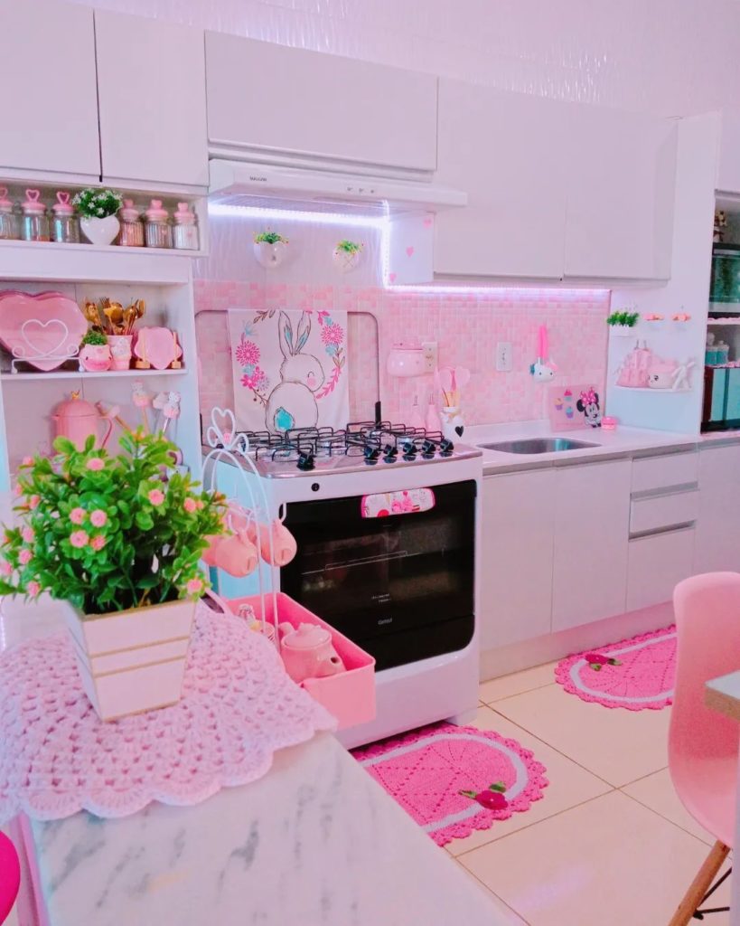 A imagem ilustra uma cozinha rosa e branca, cheia de itens decorativos em rosa pink como tapetes de crochê. 