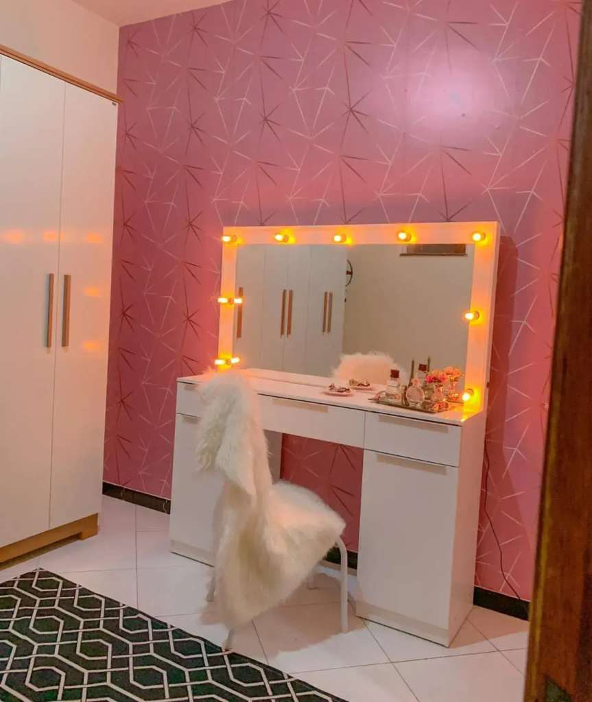 A imagem ilustra um quarto com parede rosa escuro composto por uma penteadeira camarim branca e um armário branco.
