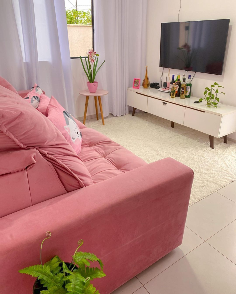 A imagem ilustra uma sala com um sofá rosa pink decorada em branco, com um hack e mesa de canto. Algumas plantas dão verde ao ambiente.