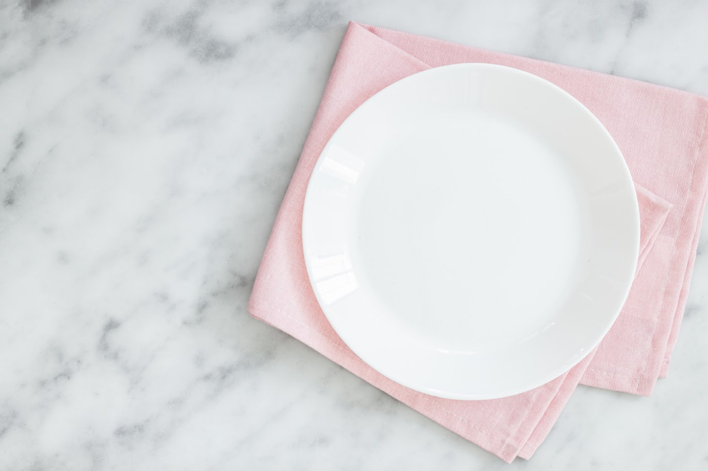 A imagem ilustra um prato branco sobre uma mesa de pedra, para ilustrar as cores que combinam com rosa, já que abaixo temos um pano de prato rosa bebê.