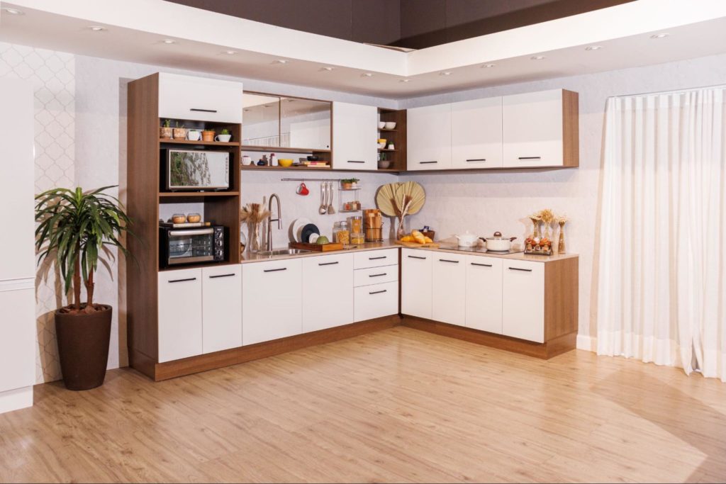 A imagem ilustra uma cozinha branca com madeira em uma sala ampla de chão de madeira. Ao seu lado há um vaso de plantas verdes.