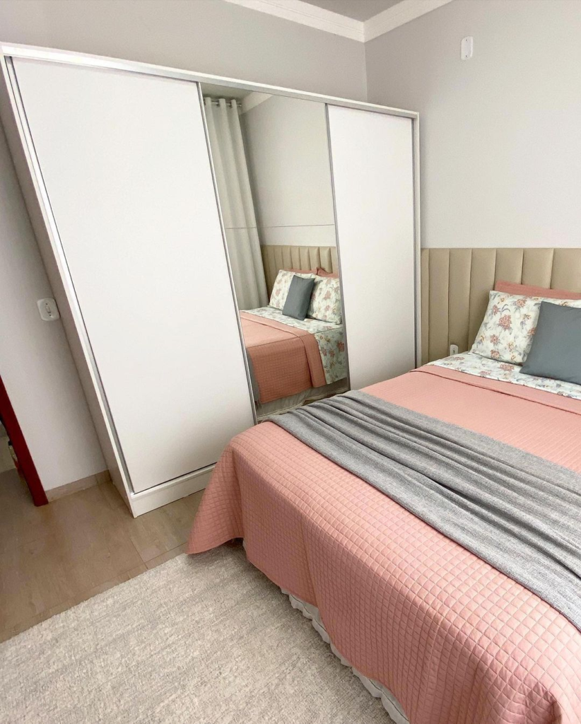 A imagem ilustra uma cama de casal com colcha rosa bebe e manta cinza ao lado de um armário branco com portas de correr e espelho no meio. 