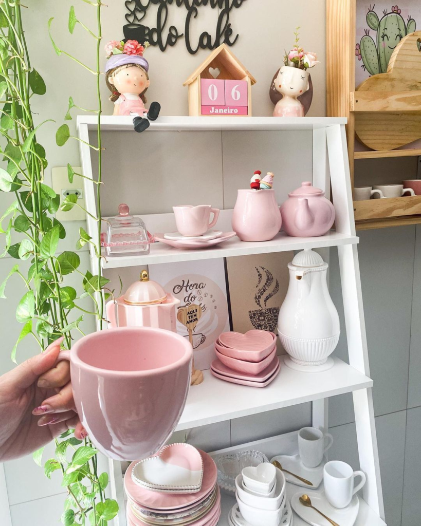 A imagem ilustra uma mão segurando uma chícara de chá rosa. Ao fundo temos uma estante escada branca repleta de louças também cor de rosas e uma folhagem verde ao seu lado.