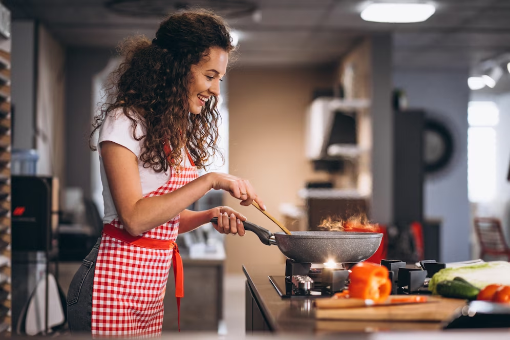 A imagem ilustra uma mulher negra de cabelos cacheados em uma cozinha gourmet pequena cozinhando em um cooktop. Ela usa um avental vermelho.