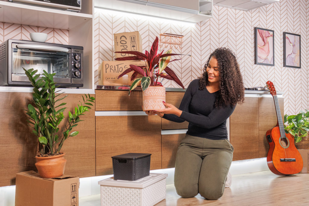 Uma mulher está abaixada em frente à sua cozinha planejada Madesa. Ela segura um vaso em suas mãos e, ao seu lado, estão caixas de organização e outro vaso de plantas. 