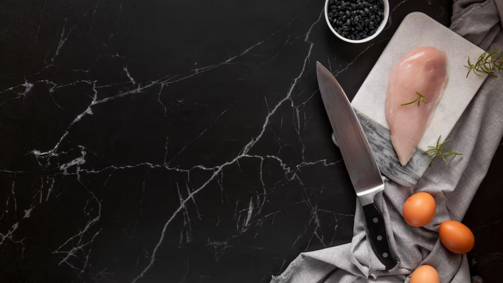 A imagem ilustra uma pedra para bancada de cozinha na versão granito, na cor preta, com uma faca de cortar carne e um pedaço de peito de frango e ovos sobre ela.